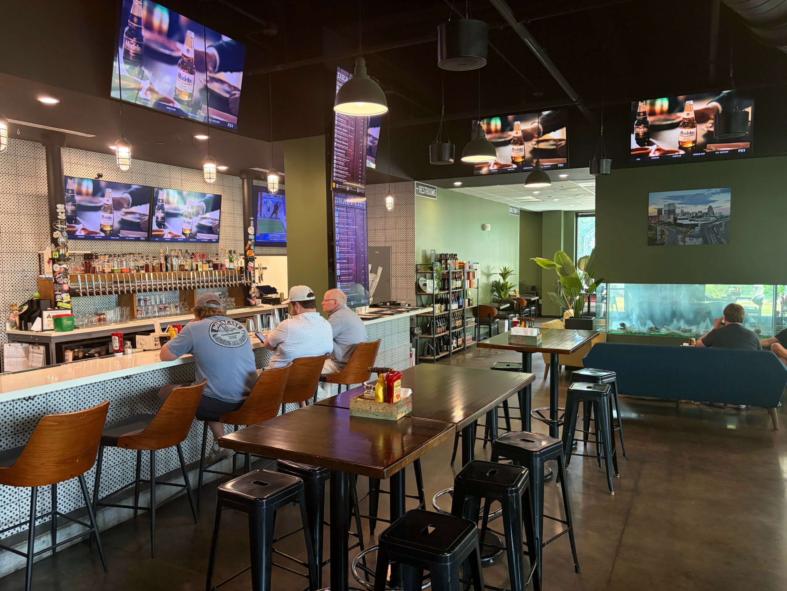Bar area with seating, multiple television screens, and a person sitting at the bar, illuminated by pendant lights.