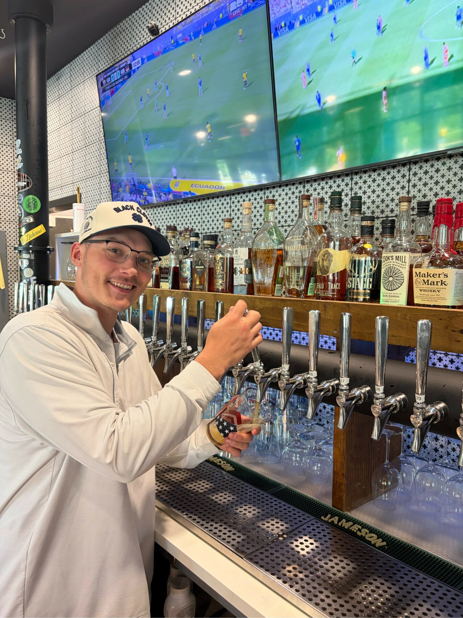 Person wearing glasses and a white jacket smiling at a bar with multiple beer taps, watching a sports game on two large screens.