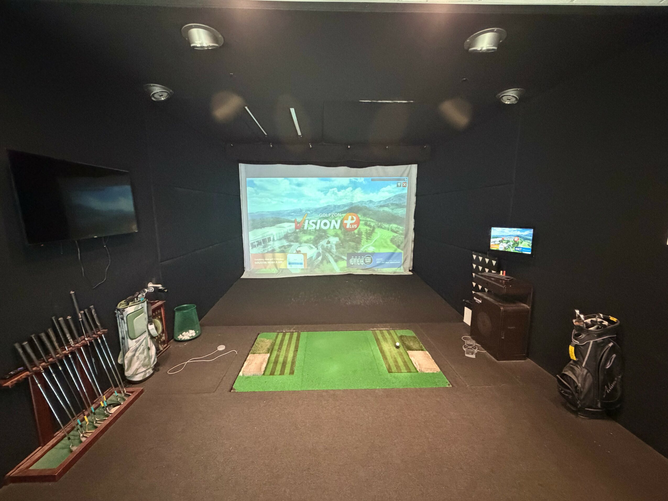 Indoor golf simulation room with screen, golf clubs, and equipment, carpeted floor, and dark walls.