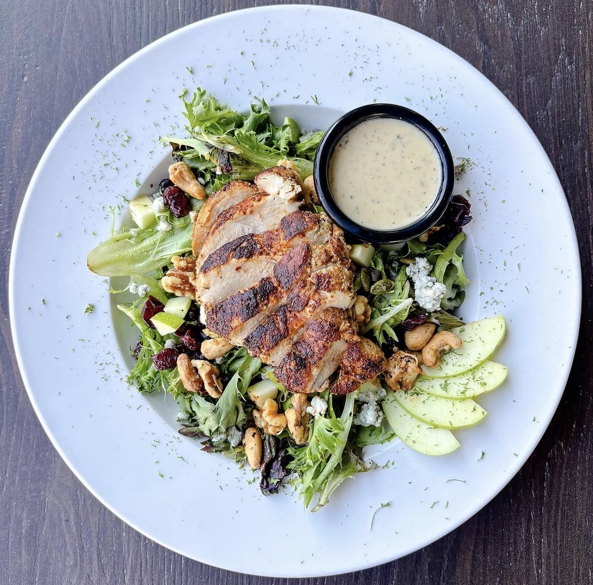 Grilled chicken breast on mixed greens with cucumber slices, nuts, and a small container of dressing on a white plate.
