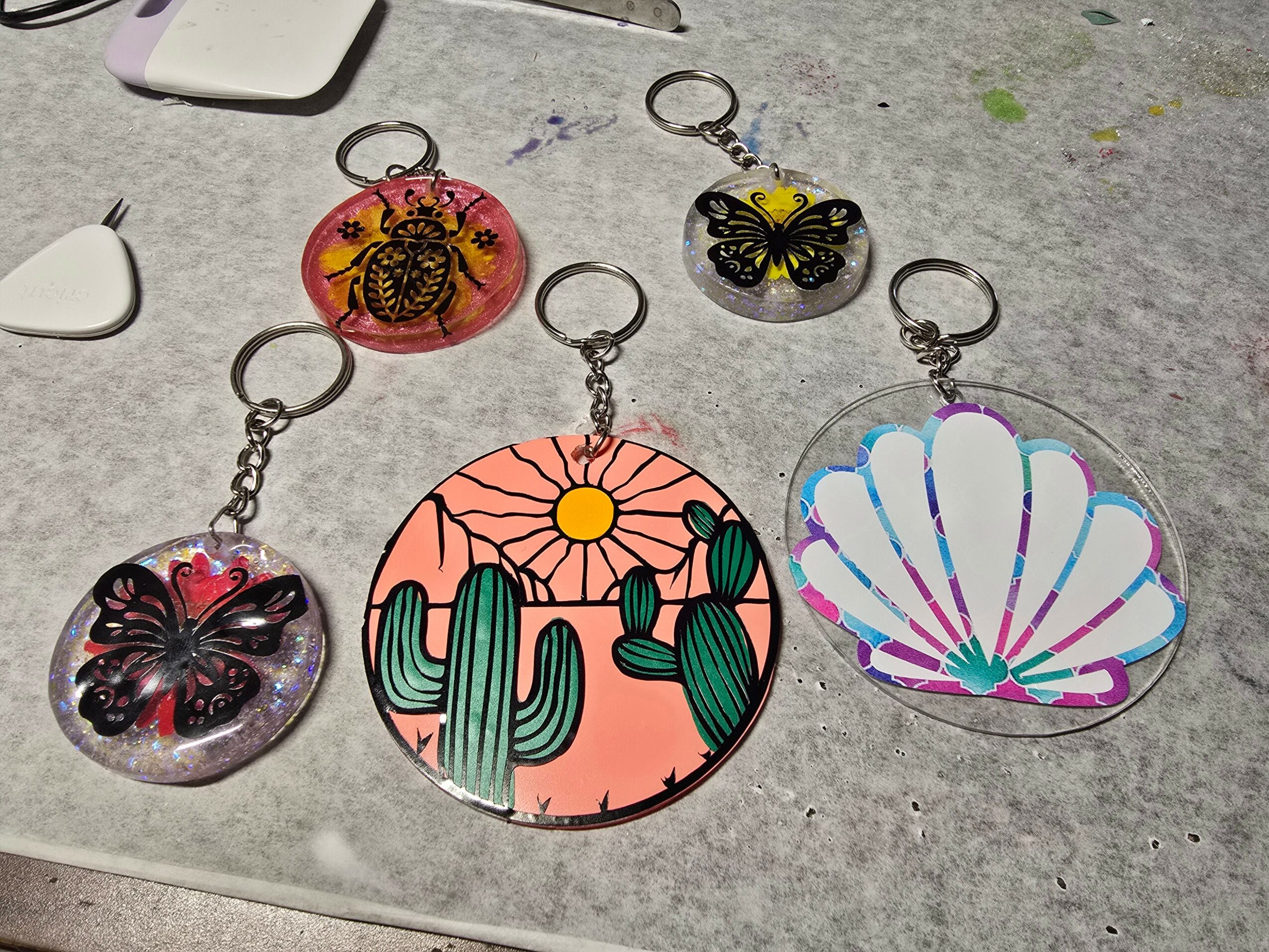 Six keychains with butterfly, cactus, and shell designs on a gray surface, some with colorful backgrounds.