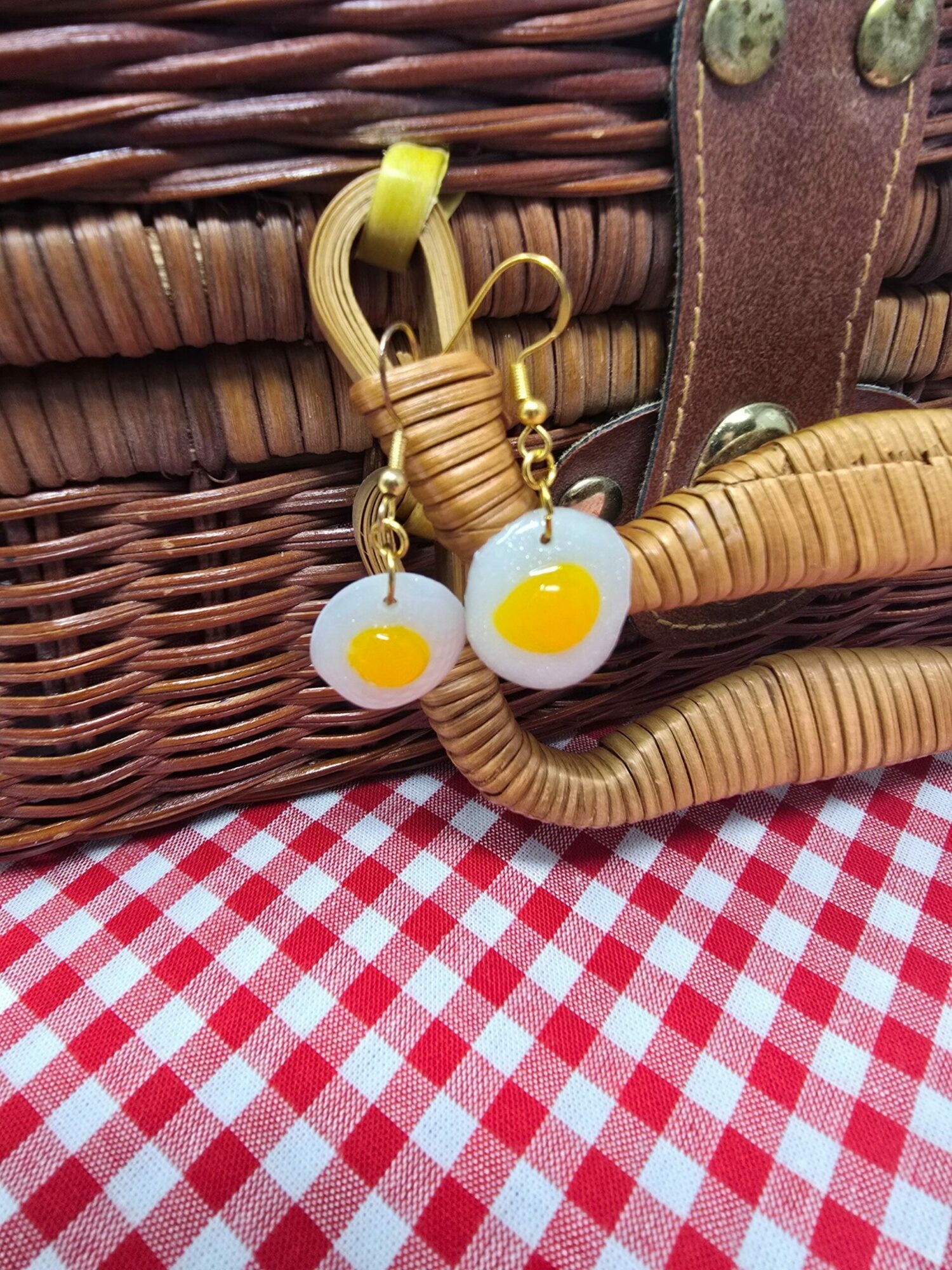 Close-up of fried egg-shaped earrings with yellow and white design, hanging on a woven bag with red and white checkered fabric background.