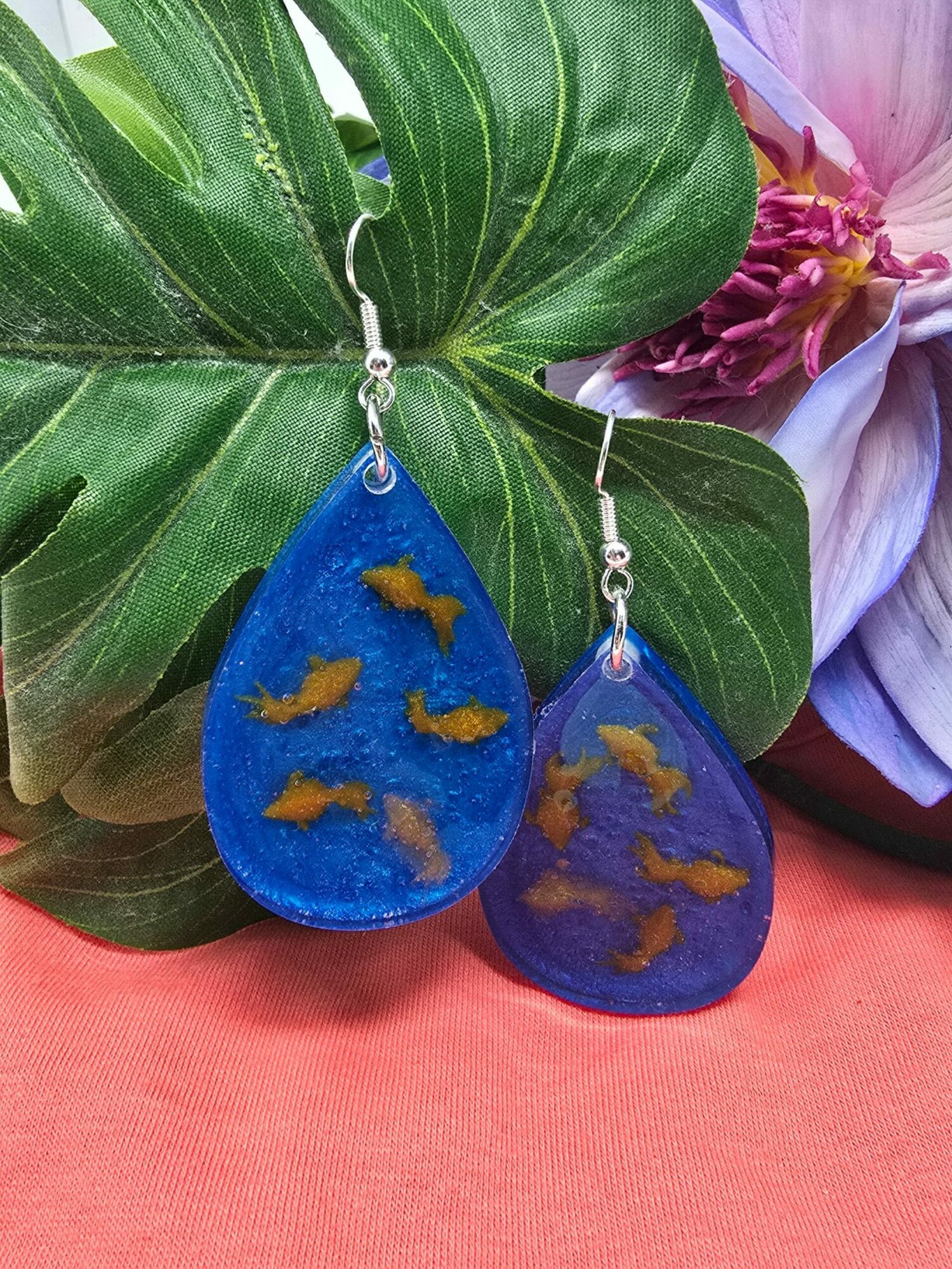 Two blue earrings with goldfish designs hang from a green leaf, with purple and pink flowers in the background.