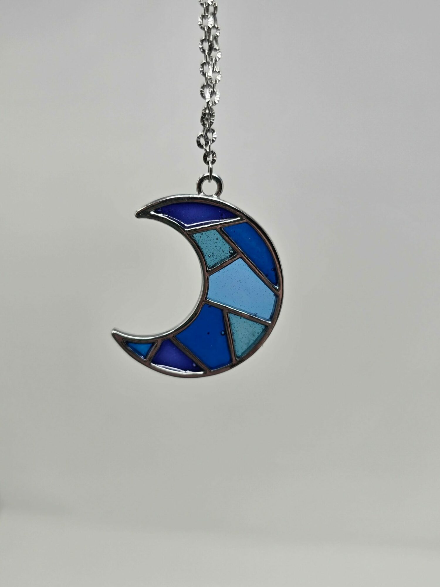 A necklace with a crescent moon pendant made of blue stained glass pieces, hanging against a plain background.
