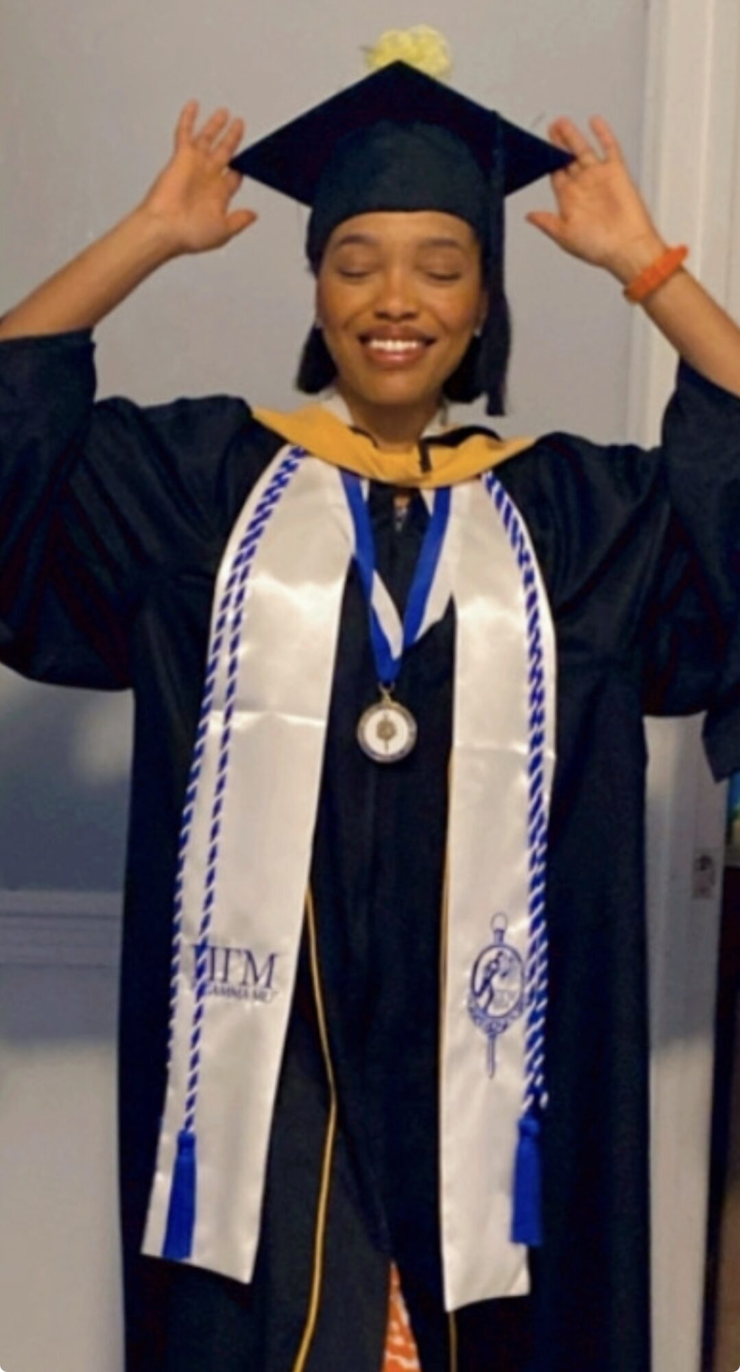 Young woman in graduation cap and gown smiling, adjusting her cap, with honor cords and medals, indoors.