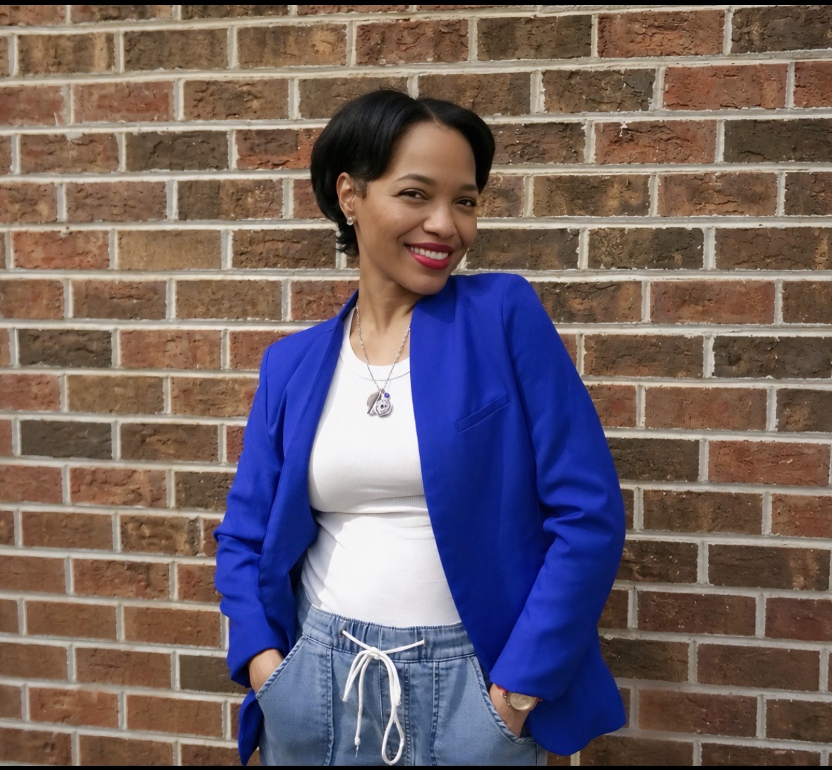 Woman with short black hair smiling, wearing a blue jacket and white top, standing against a brick wall.
