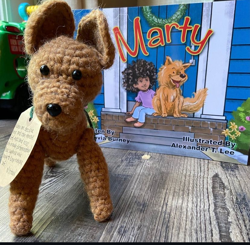 A crocheted dog plush toy with black eyes and nose, standing on a wooden surface, with a colorful children's book in the background.