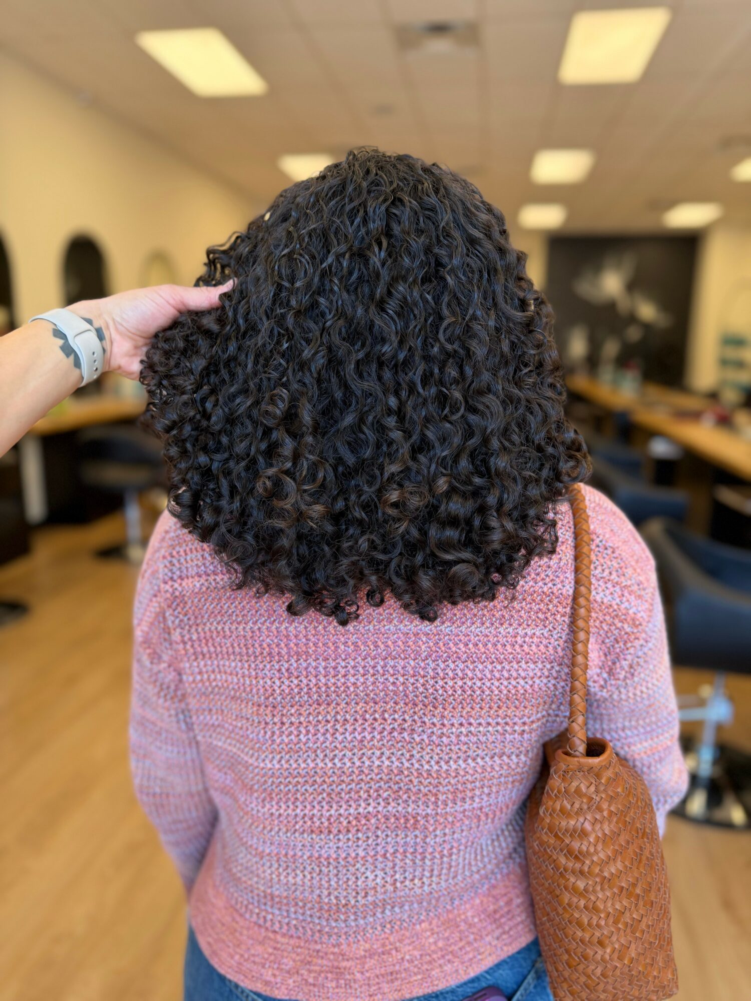 Back view of a person with curly hair wearing a pink sweater and carrying a brown woven bag in a room with tables and chairs