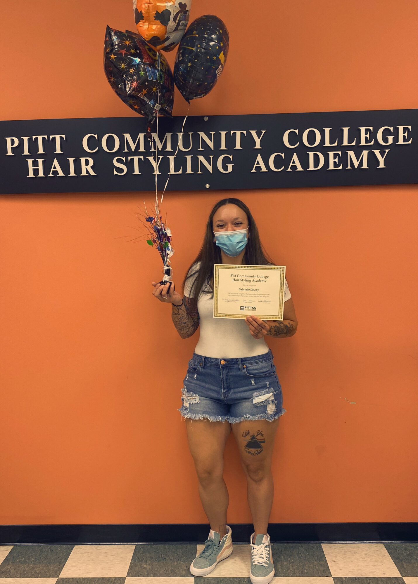 Person holding a certificate and balloons, standing in front of an orange wall with a sign for Pitt Community College Hair Styling Academy, wearing a mask, white top, and denim shorts.