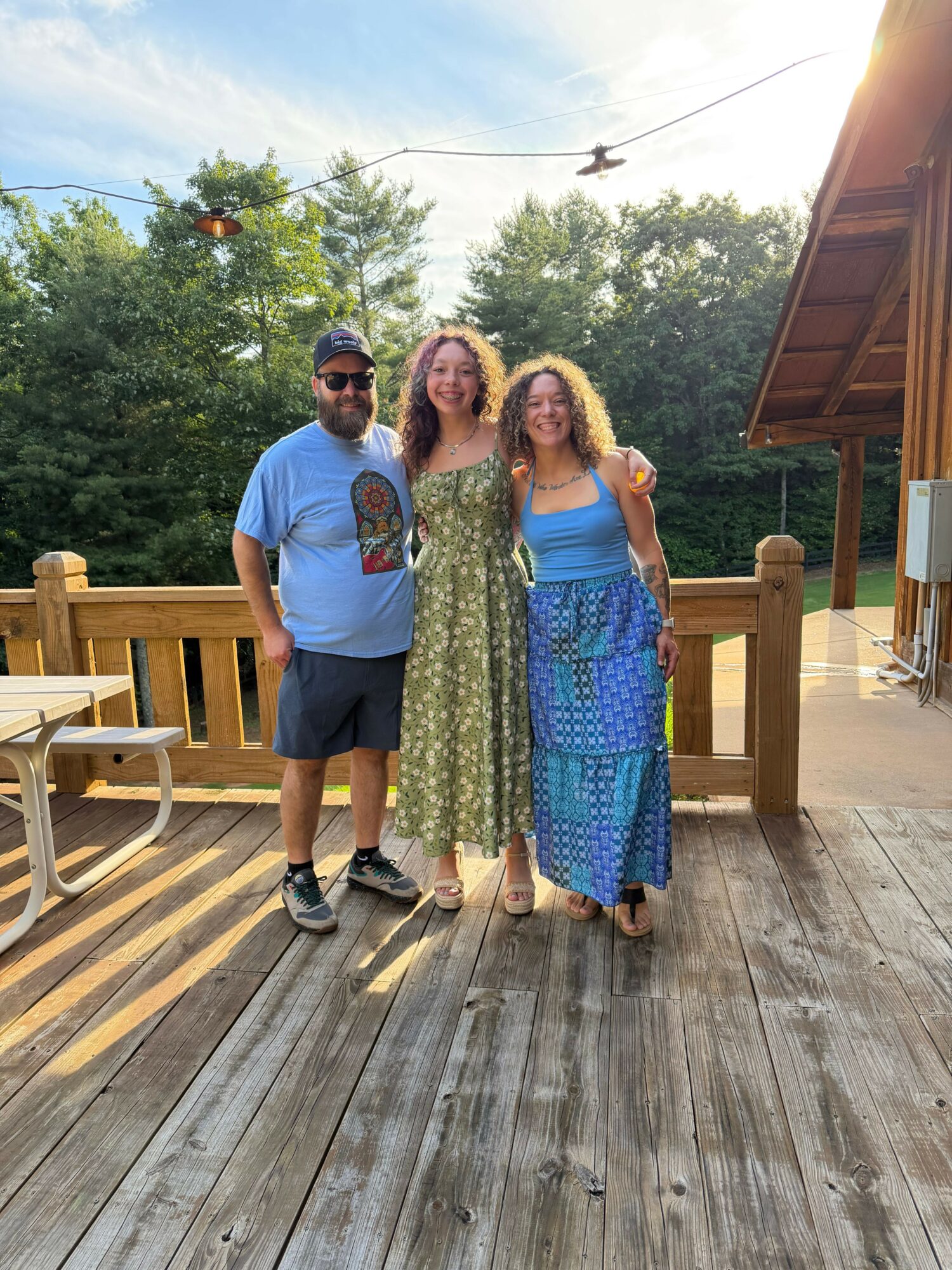 Two women stand on a wooden porch with trees and sky in the background. One woman wears a blue dress, the other a blue shirt and shorts.