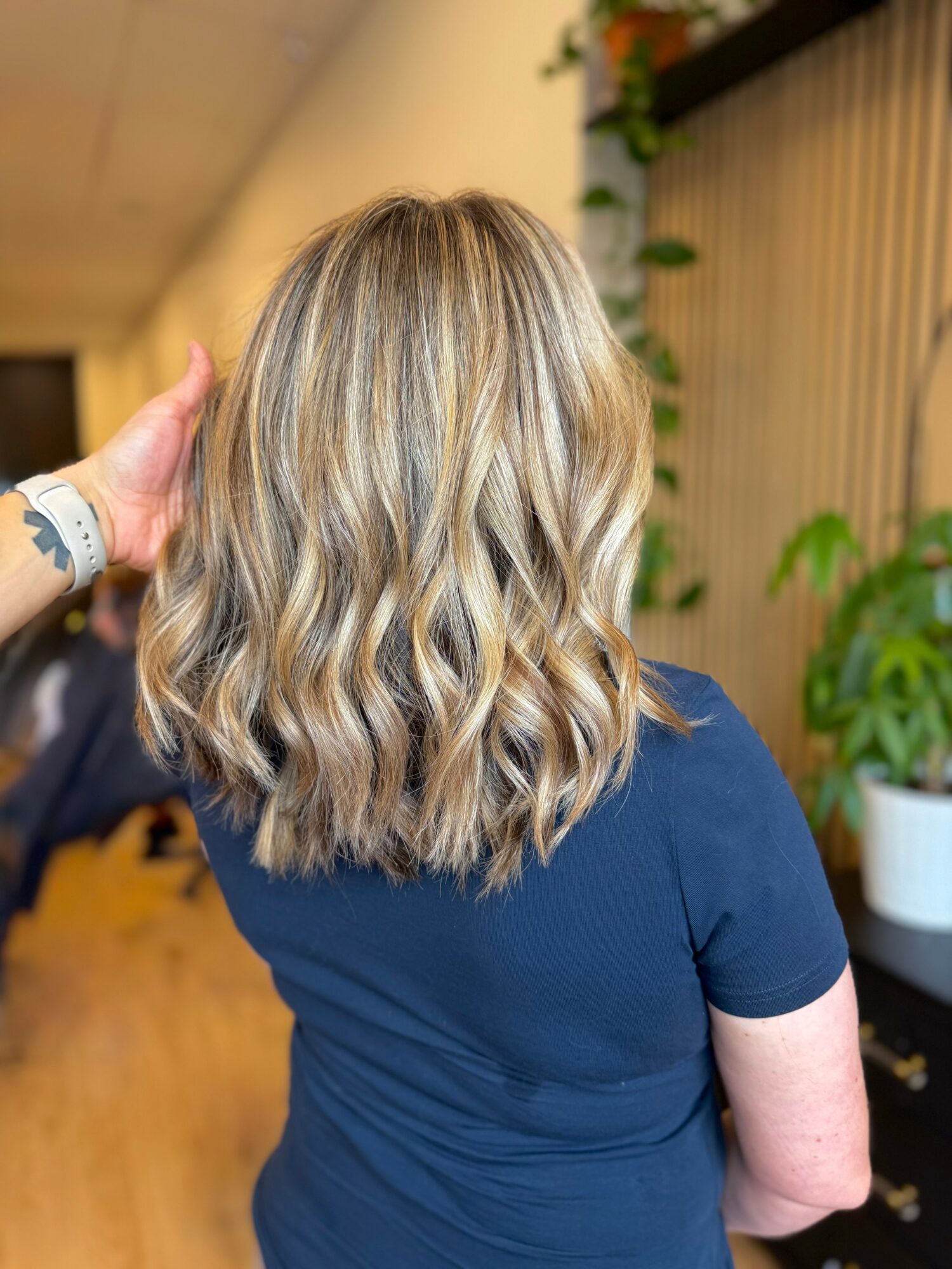 Back view of a woman with shoulder-length wavy blonde hair, wearing a navy blue shirt, in an indoor setting with plants.