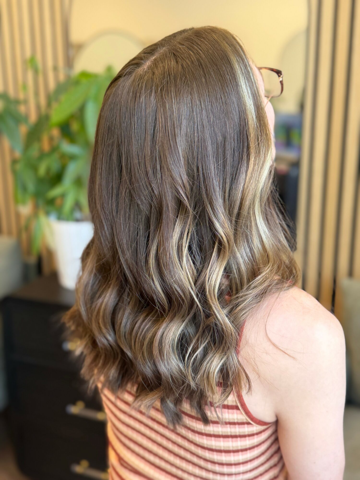 Back view of a woman with long, wavy, light brown hair, wearing a striped sleeveless top, in an indoor setting.