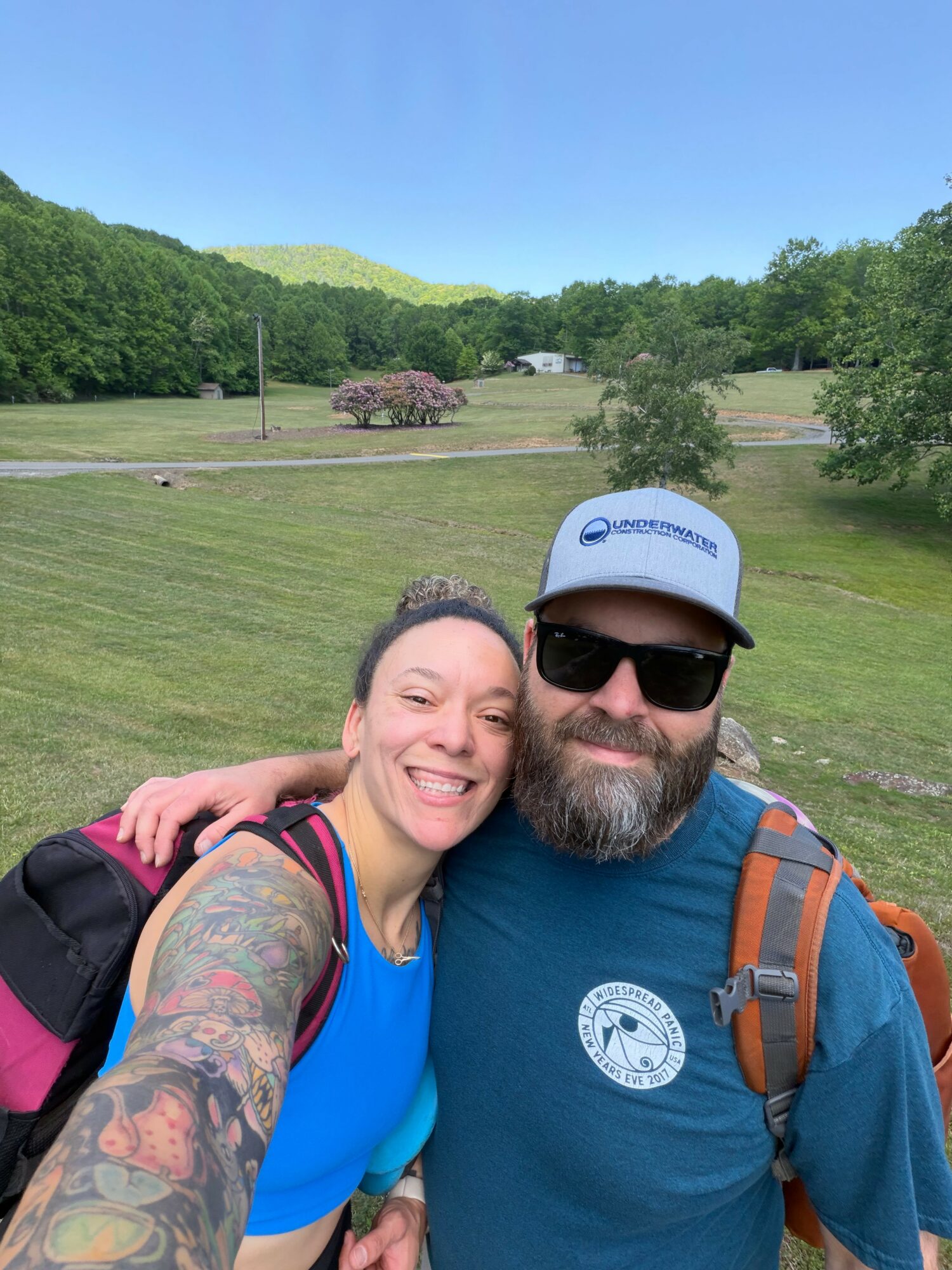 Two people smiling outdoors with green hills and trees in the background, one wearing sunglasses and a cap, the other with tattoos.