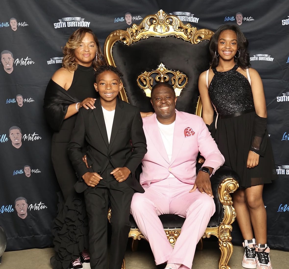 Four people, one seated on a gold and black throne, three standing around, all smiling, black backdrop with logos, formal attire.