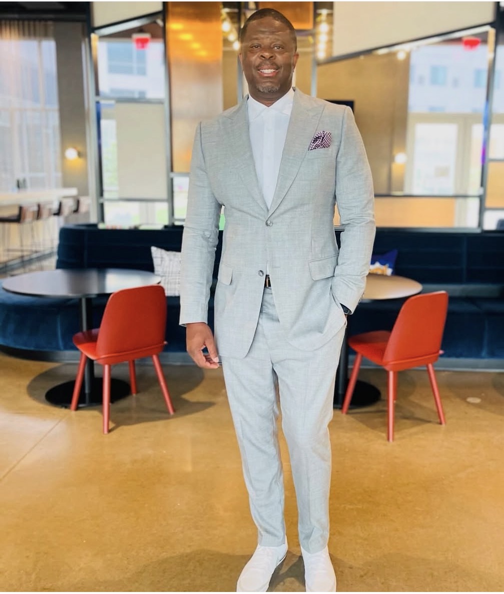 Man in light gray suit standing in a modern restaurant with red chairs and large windows.