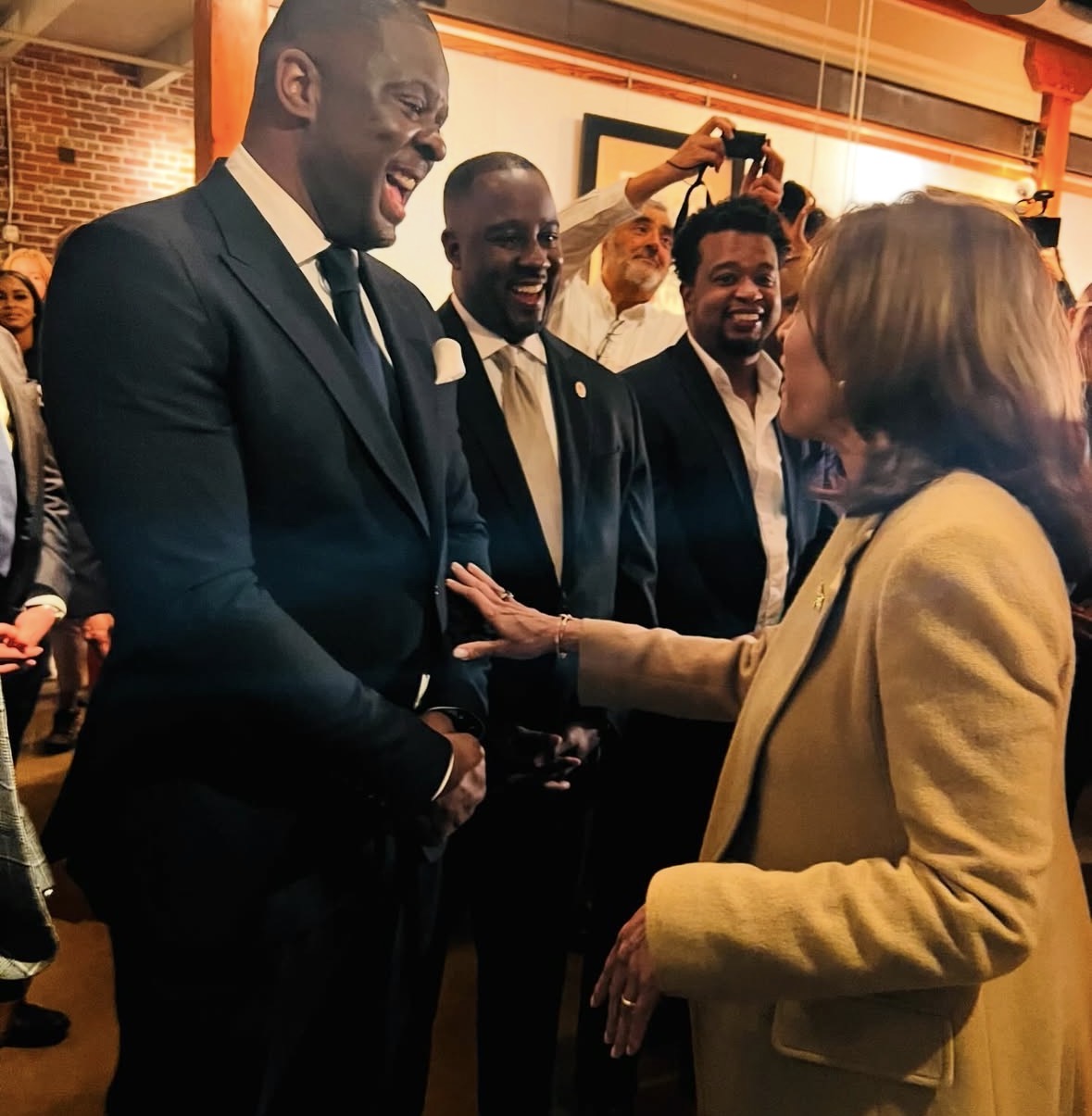 Group of people smiling and talking indoors, woman in yellow jacket engaging with man in suit, warm lighting.