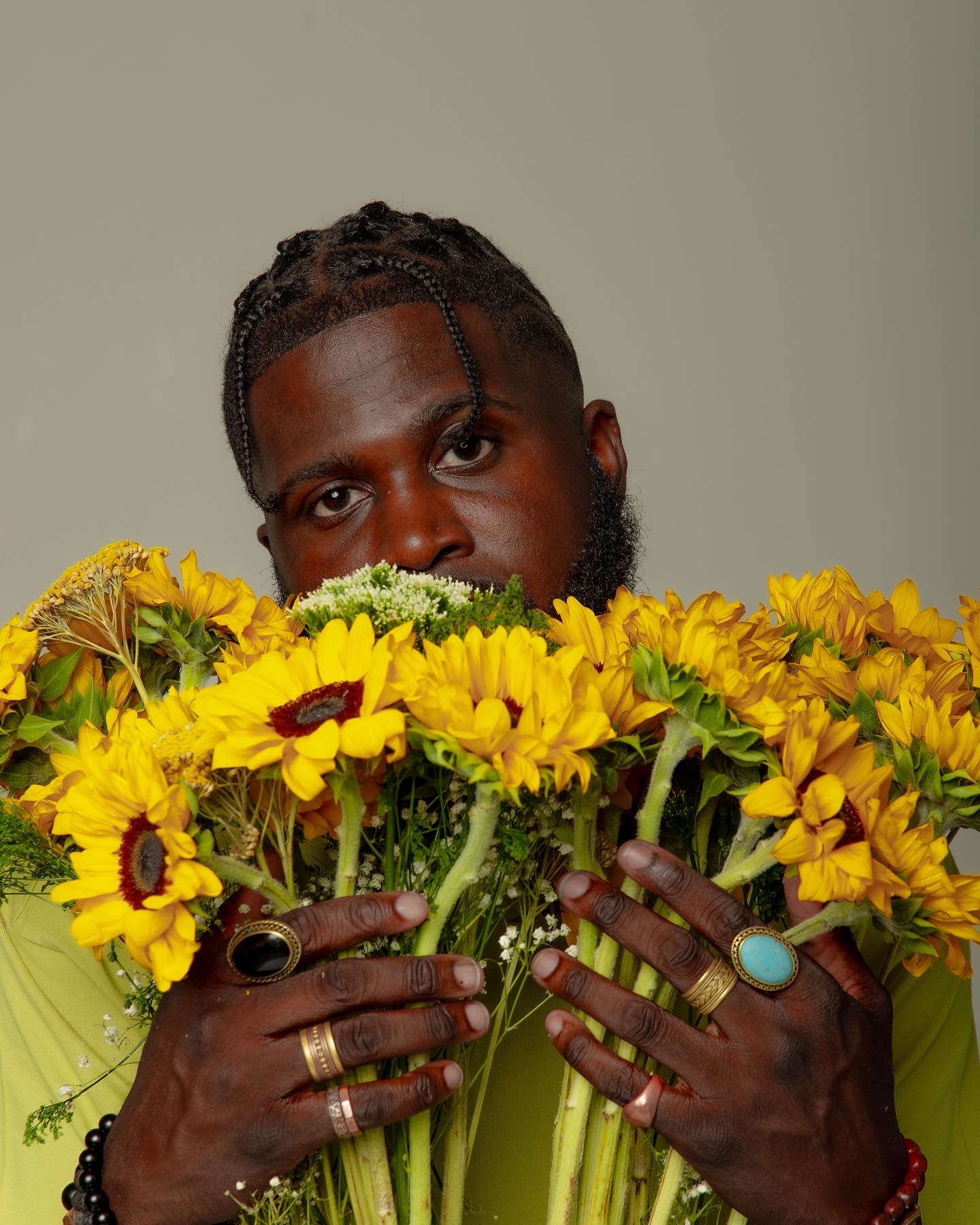 Person with braided hair and beard holding a large bouquet of yellow flowers, wearing rings and a bracelet, against a plain background.