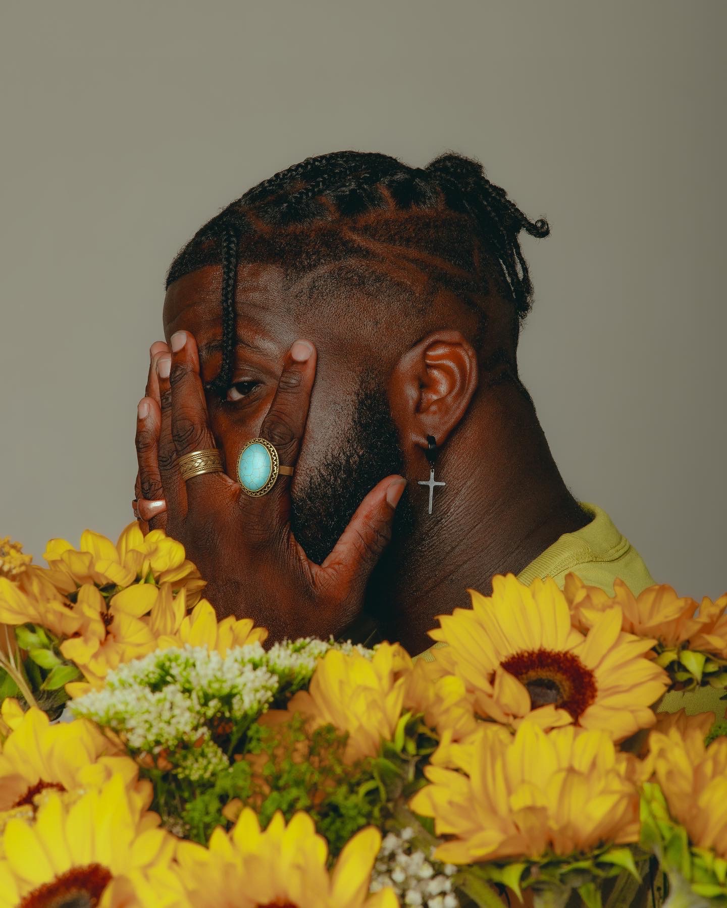 Person with styled hair and jewelry, covering face with hand, surrounded by yellow flowers.
