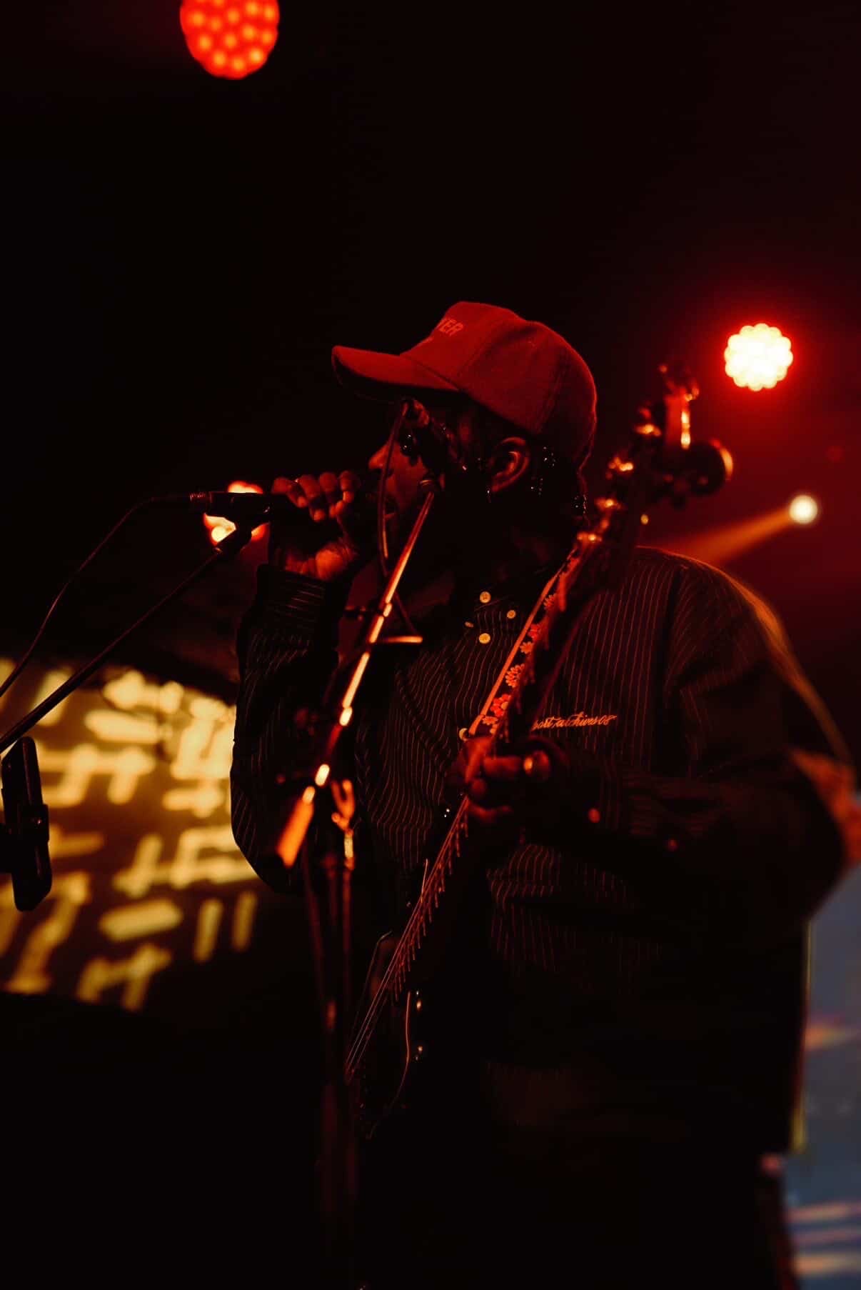 Person wearing a cap and jacket, holding a microphone and guitar on stage with red and orange lights.