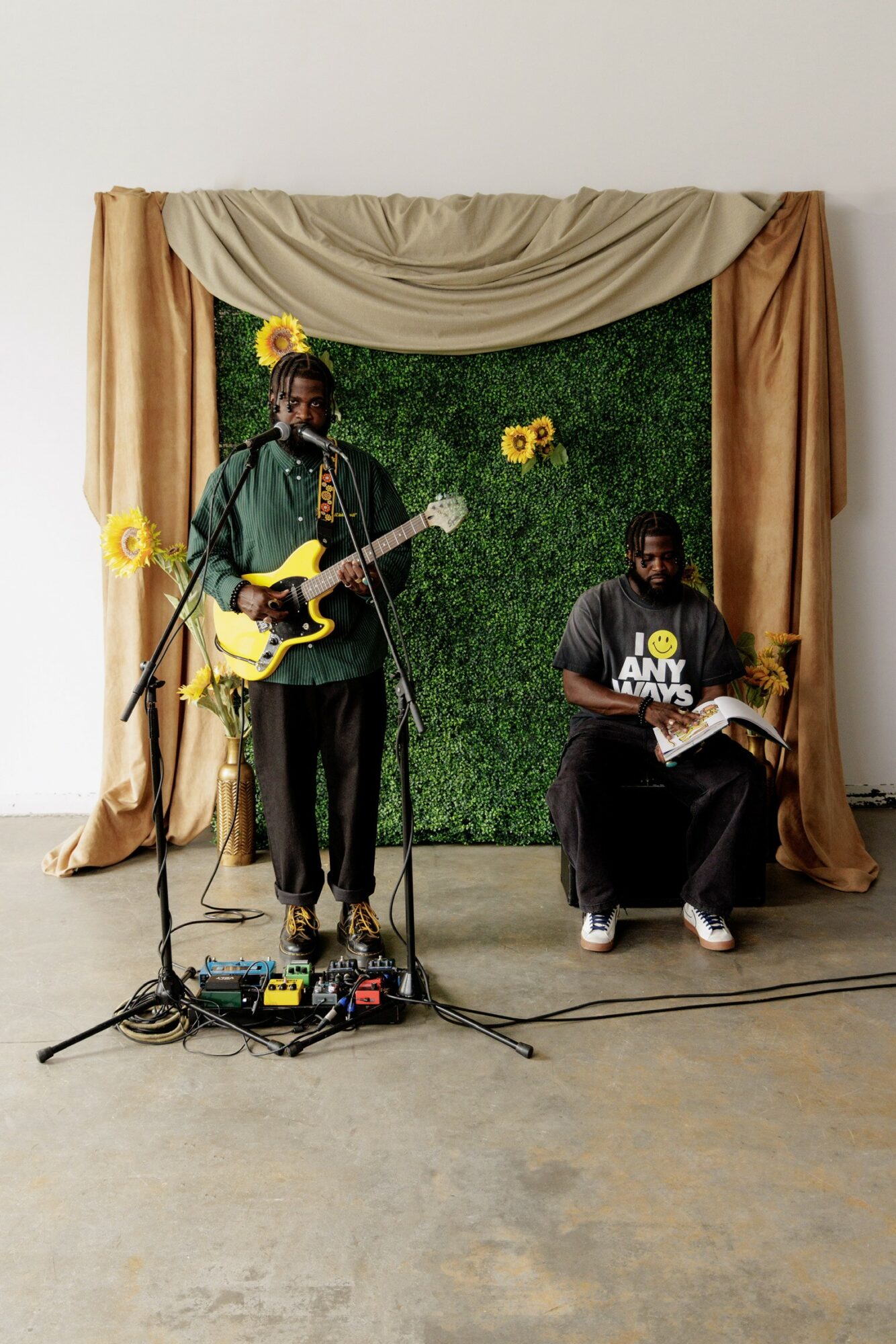 Two musicians perform on stage with a green backdrop and yellow flowers, one singing and playing guitar, the other reading music.