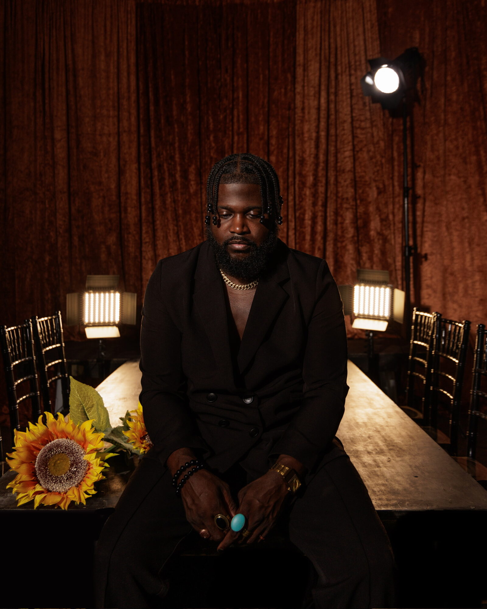 Man with dreadlocks and beard sitting at a table with sunflowers, in a wood-paneled room, with lighting equipment visible.