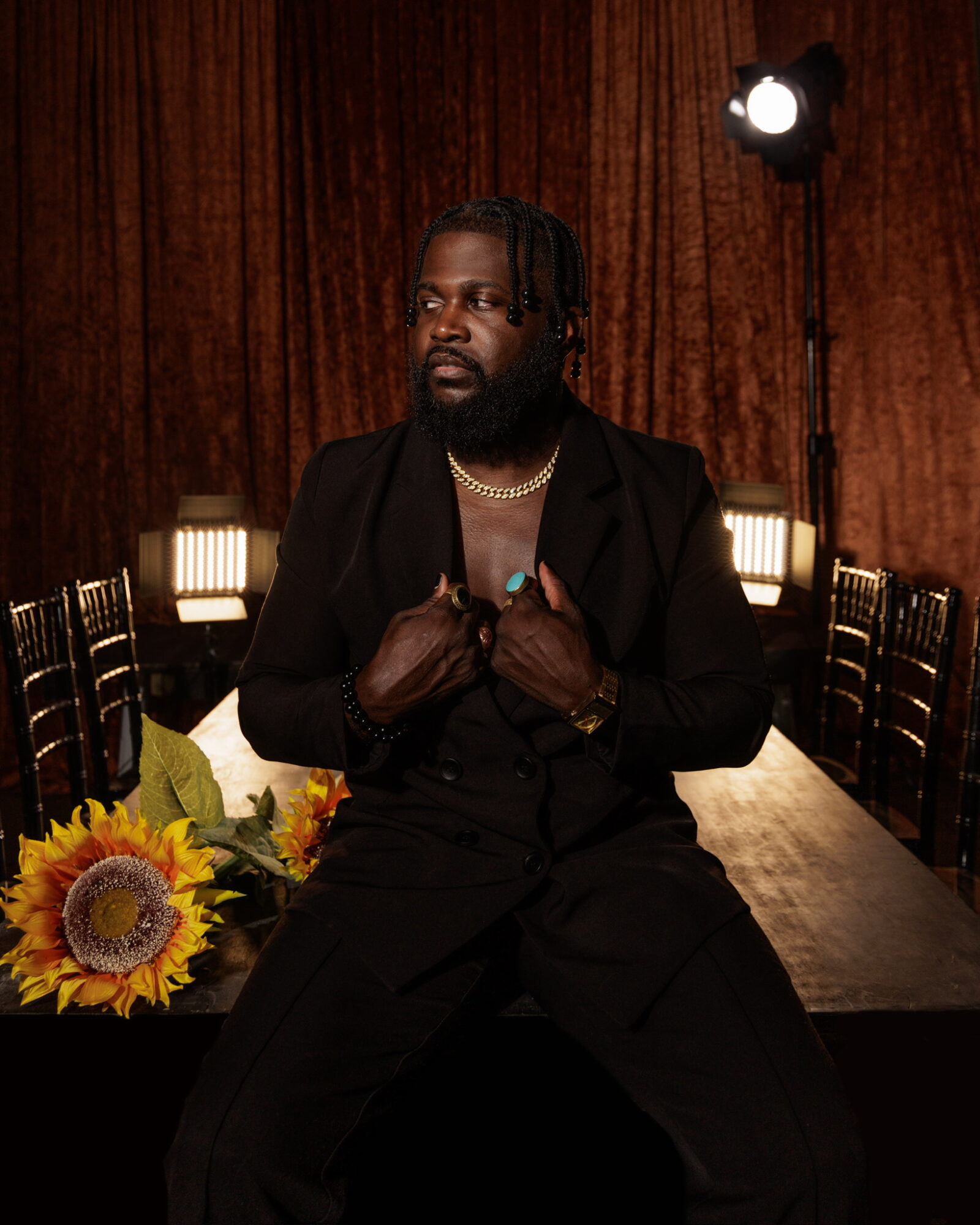 Man with dreadlocks and beard wearing a black suit, sitting at a table with sunflower bouquet, in a dimly lit room.