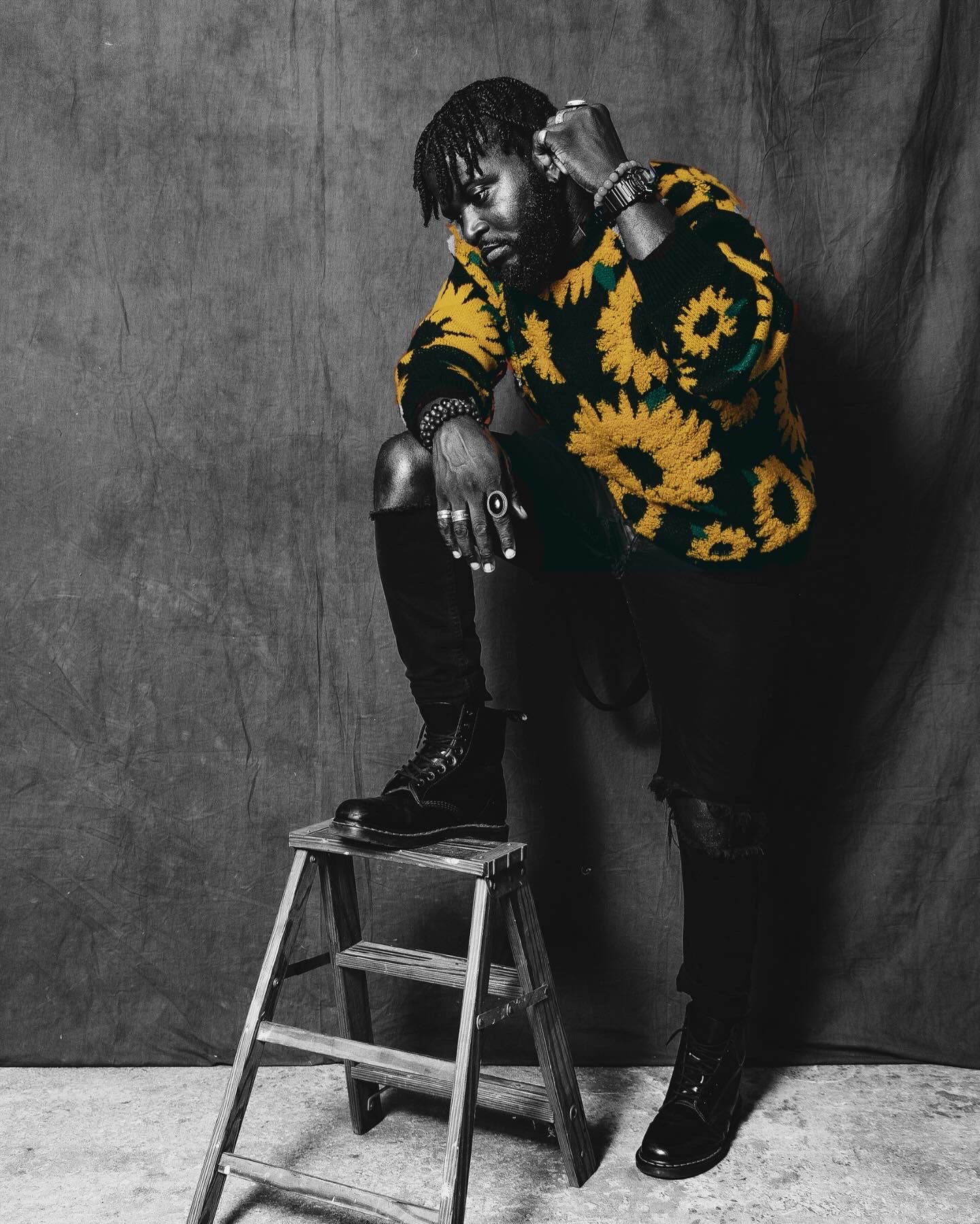 Person with dreadlocks wearing a sunflower-patterned sweater, sitting on a stool, with a dark backdrop behind them.