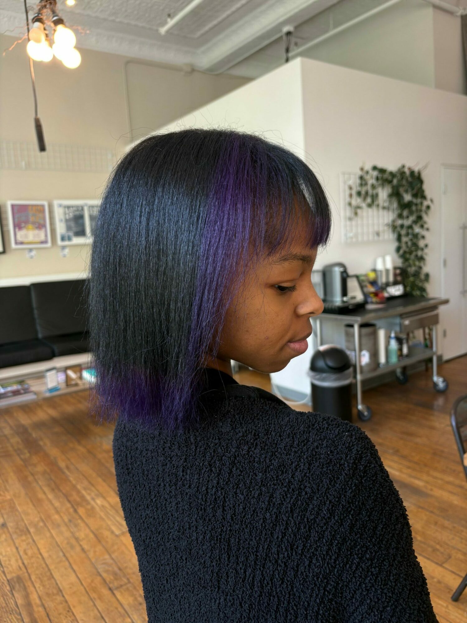 Side view of a woman with straight, shoulder-length hair, in a room with wooden floor and kitchen background.