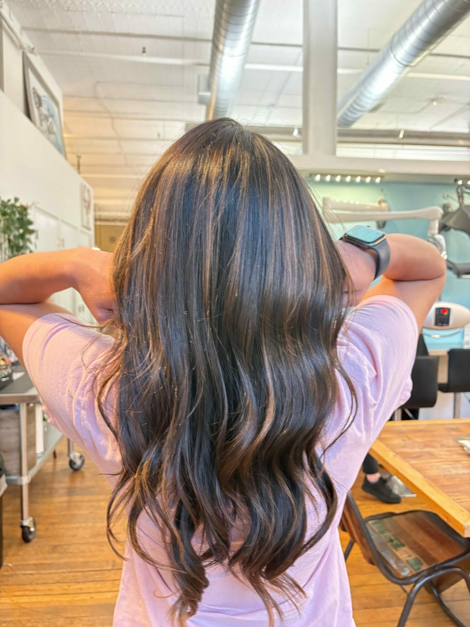 Person with long, wavy hair sitting at a desk in a room with wooden flooring and large windows.
