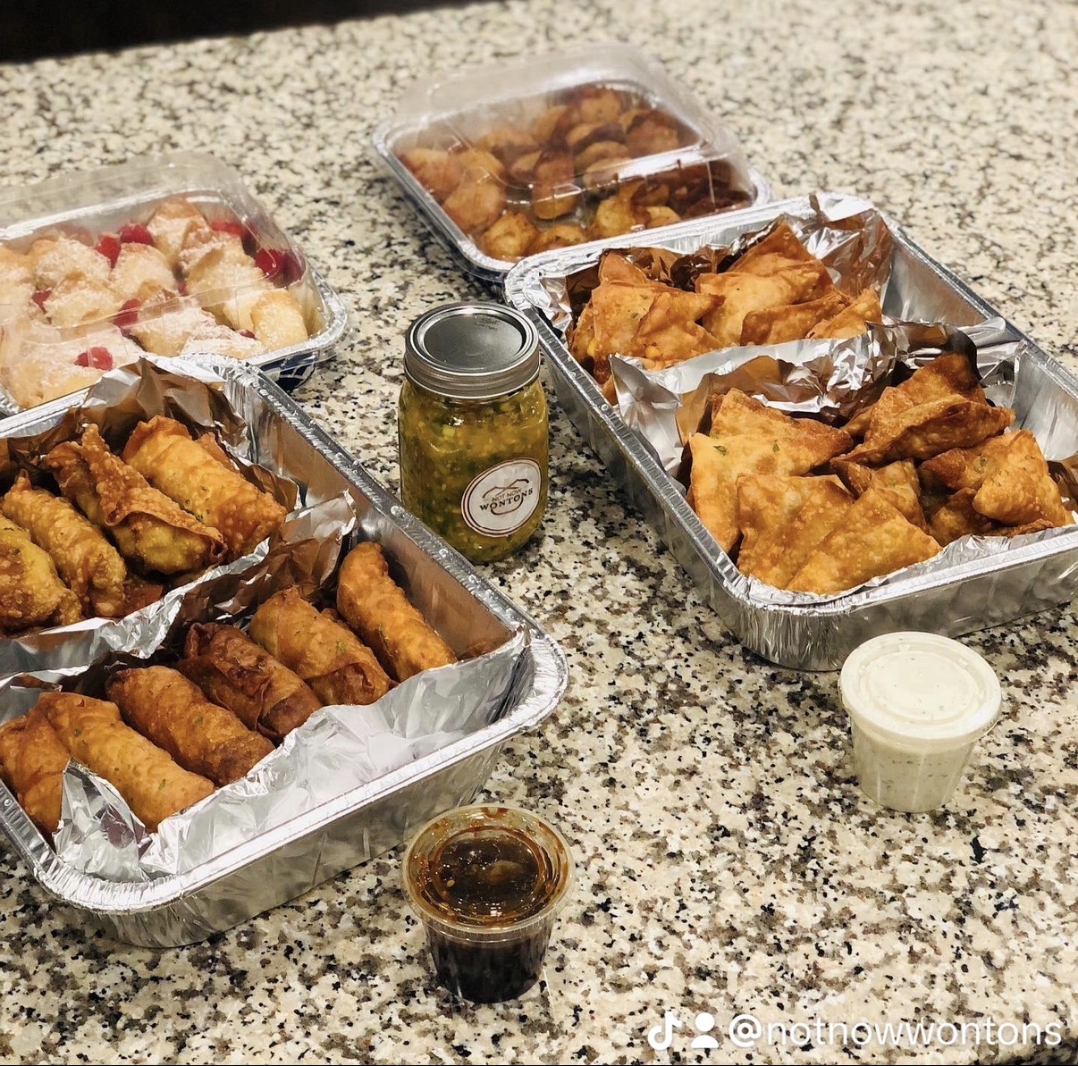 Assorted fried foods, sauces, and pickles on a speckled countertop, in aluminum trays and small jars.