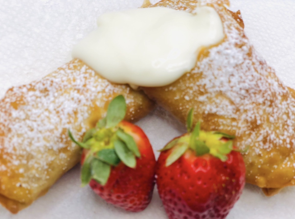 Two fried pastries dusted with powdered sugar, topped with cream, accompanied by two strawberries with green leaves.