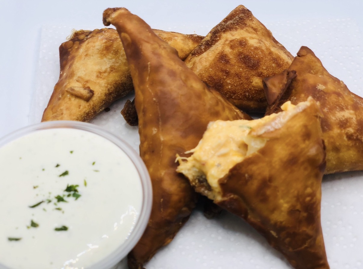 Fried samosas with a side of white dipping sauce garnished with herbs, on a white surface.