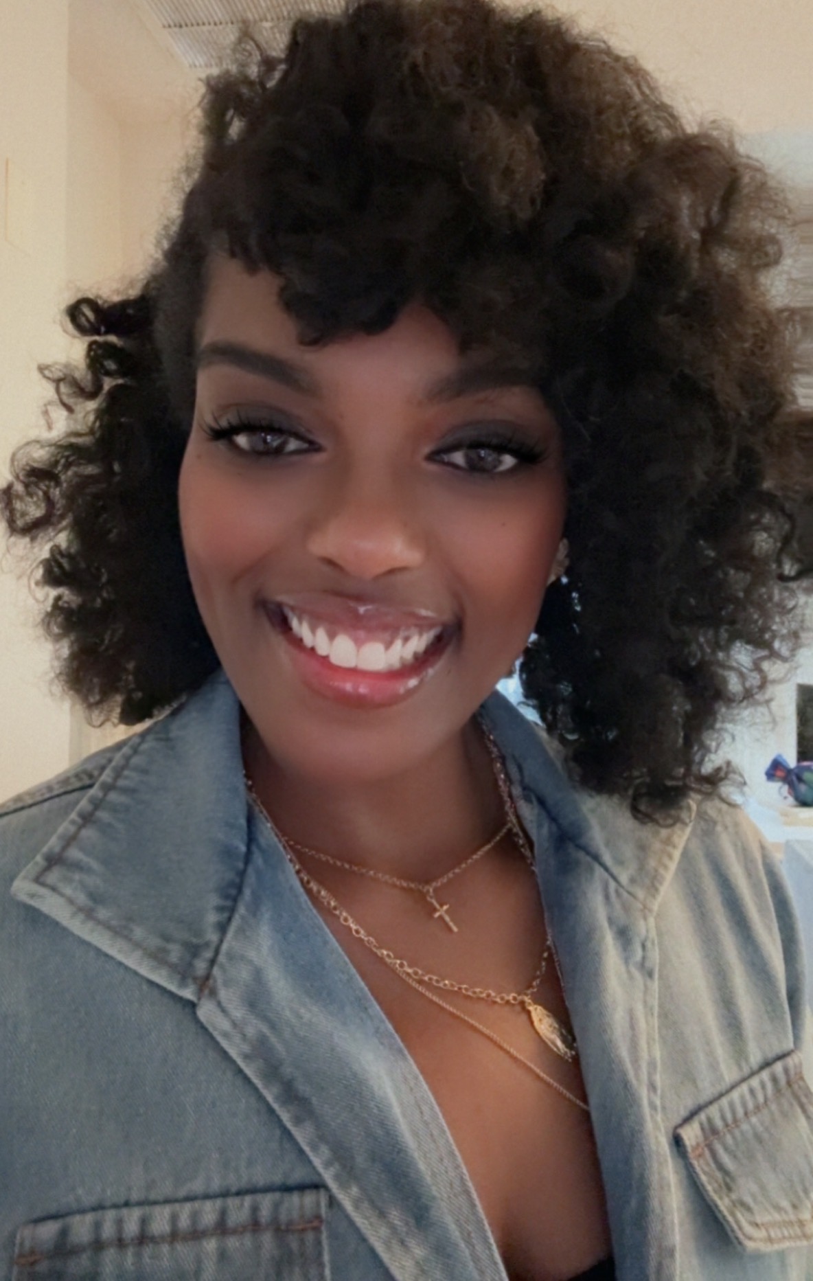 Smiling woman with curly hair wearing a denim jacket and layered necklaces.