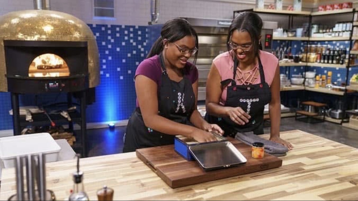 Two women wearing aprons prepare food in a kitchen with a pizza oven and shelves in the background.