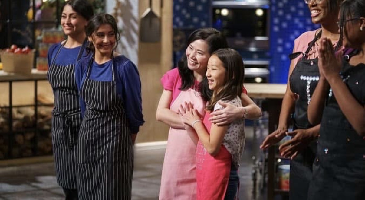 Group of women smiling and hugging in a kitchen setting, some wearing aprons, celebrating together.