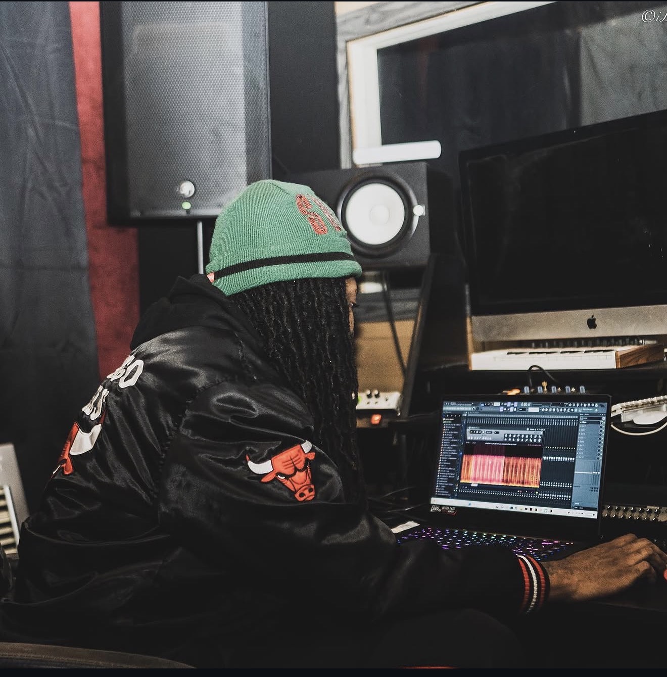 Person with dreadlocks wearing a green beanie and black jacket working on a music production laptop in a studio.