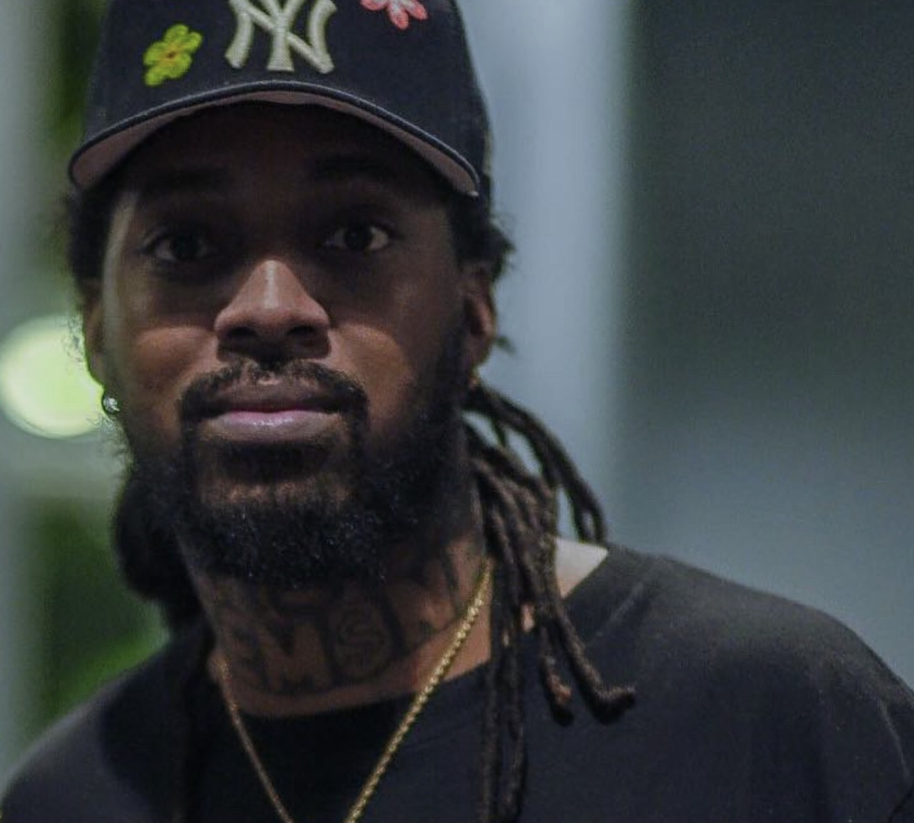 Portrait of a man with a beard, dreadlocks, wearing a cap with floral embroidery, and a black shirt.