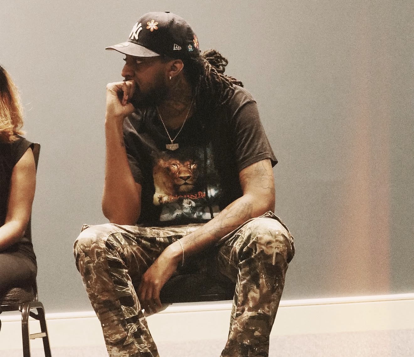Person with long dreadlocks, wearing a cap, t-shirt, and camouflage pants, sitting on a chair against a plain wall.