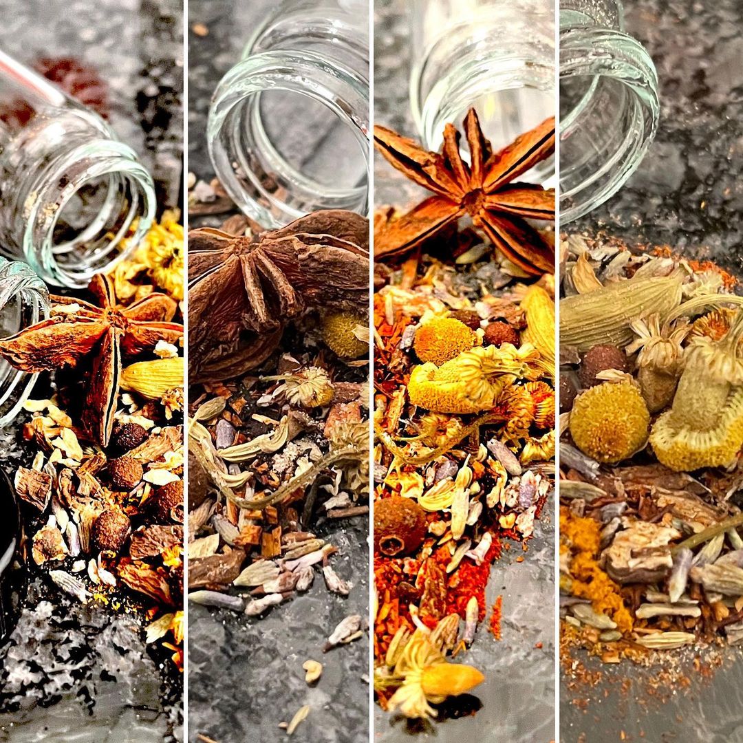 Spilled dried herbs and spices on a dark surface with a glass jar, star anise, and various dried flowers.