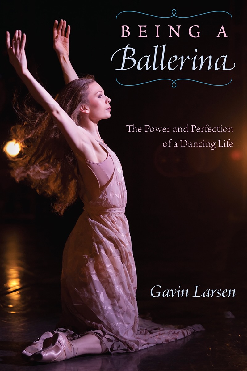 A ballerina kneels on stage with arms raised, wearing a pink dress, with dark background and stage lighting.
