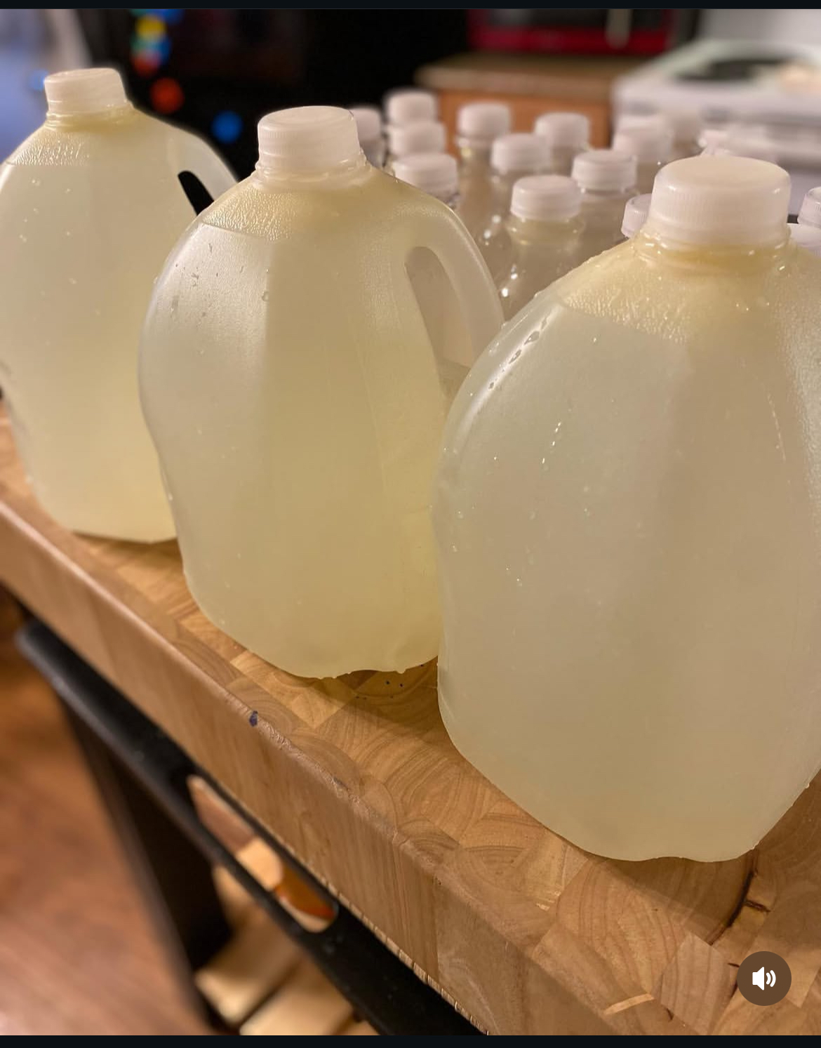 Three large plastic jugs filled with liquid on a wooden surface, with smaller bottles in the background.