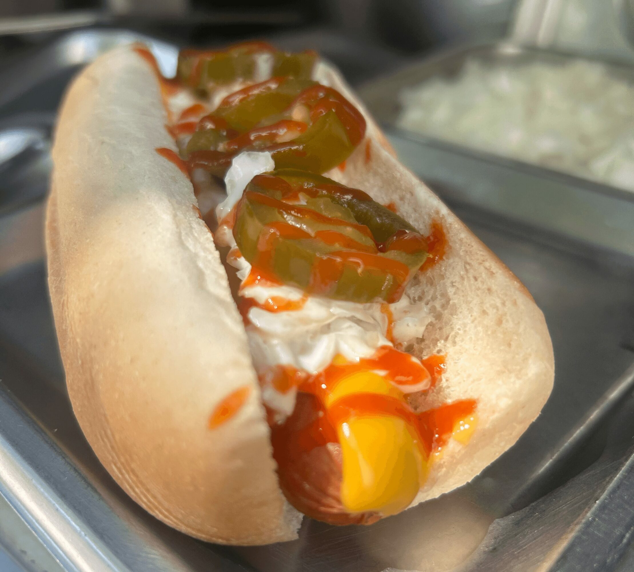 Hot dog in a bun topped with sliced pickles, ketchup, mustard, and mayonnaise, on a metal tray.