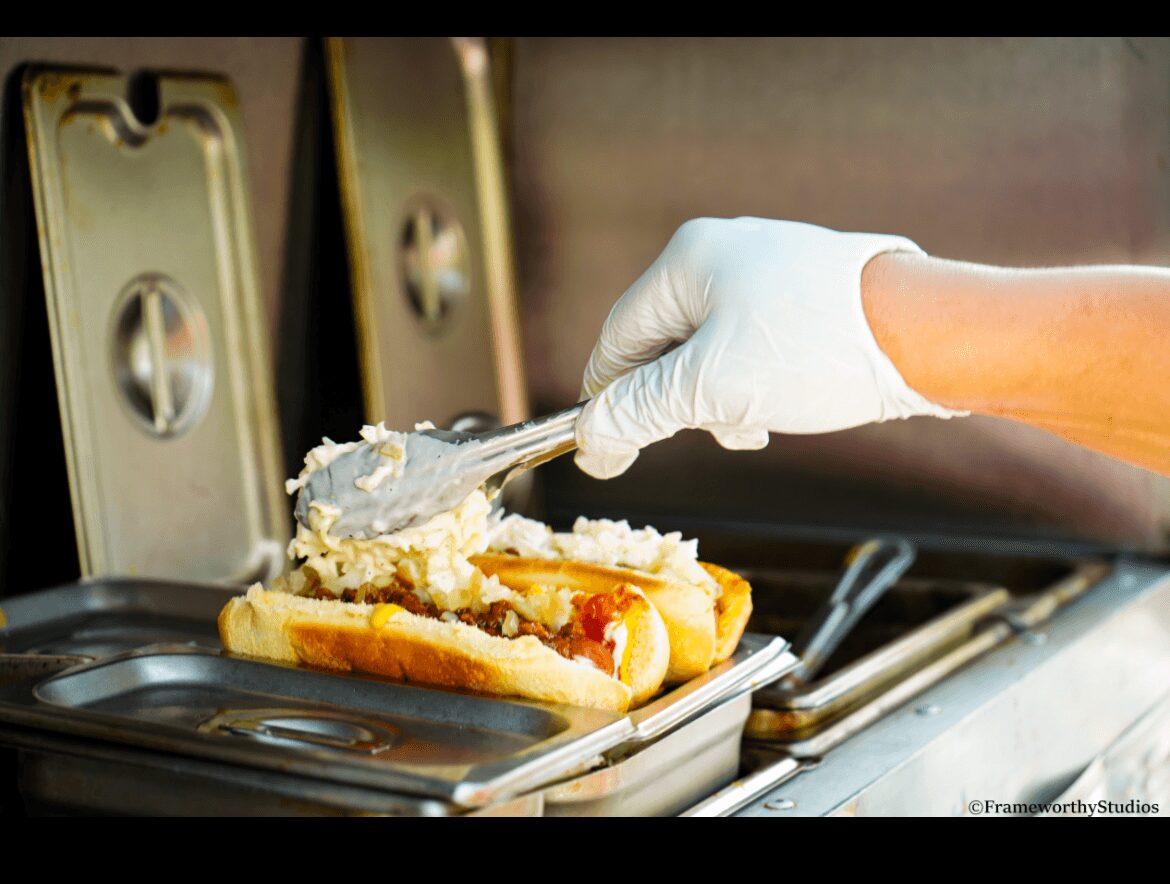 Person wearing a glove is adding toppings to a hot dog with a spoon, on a food service counter.