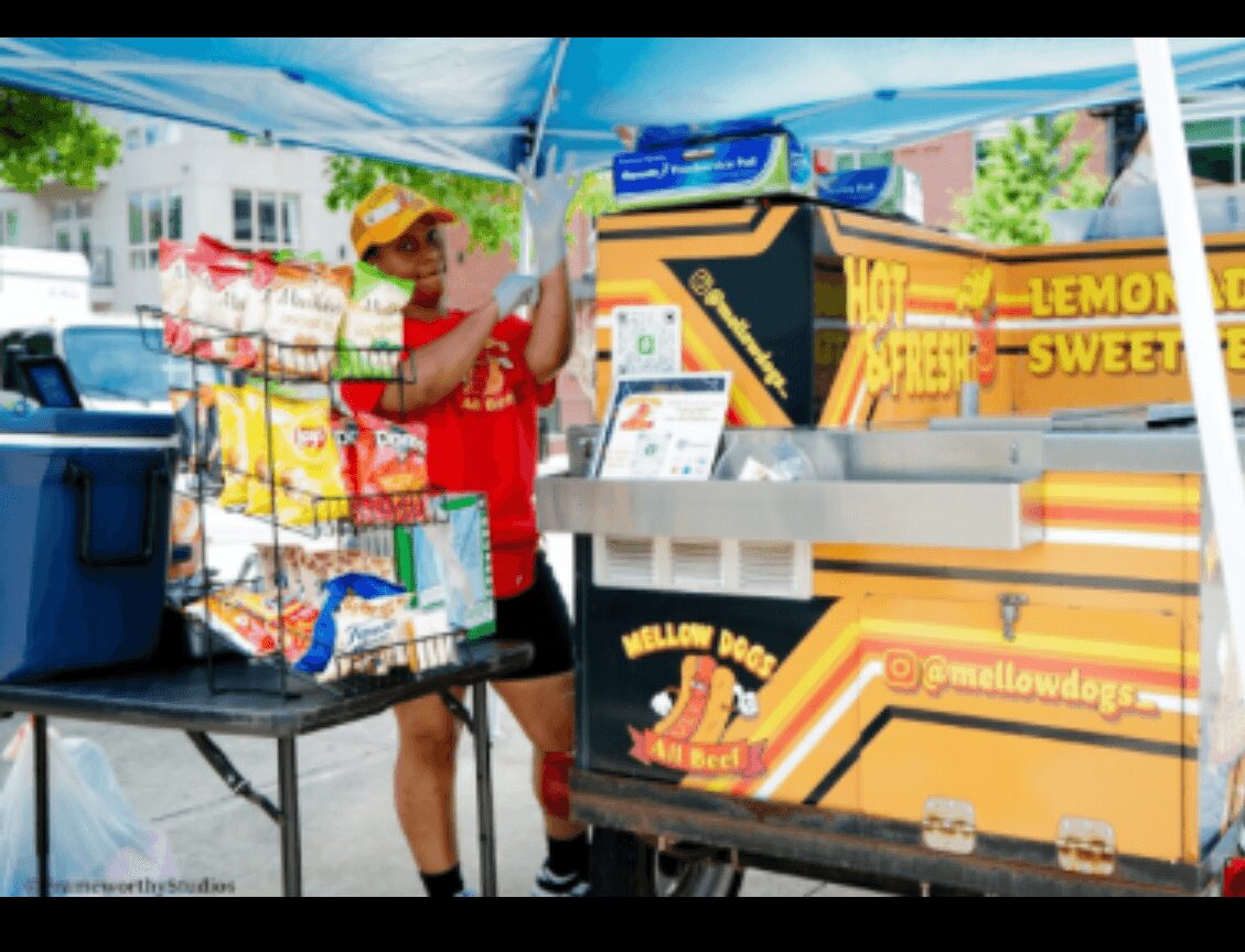 Person selling snacks at a street stand with colorful boxes and a blue canopy, outdoors in a sunny area.