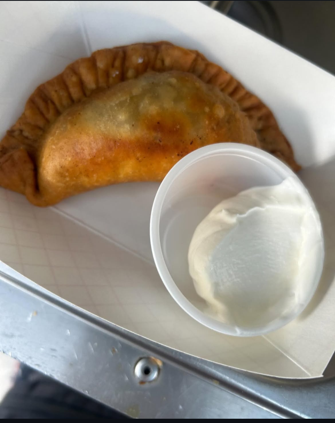 Fried empanada with a cup of white dipping sauce inside a white container, in a paper-lined box.