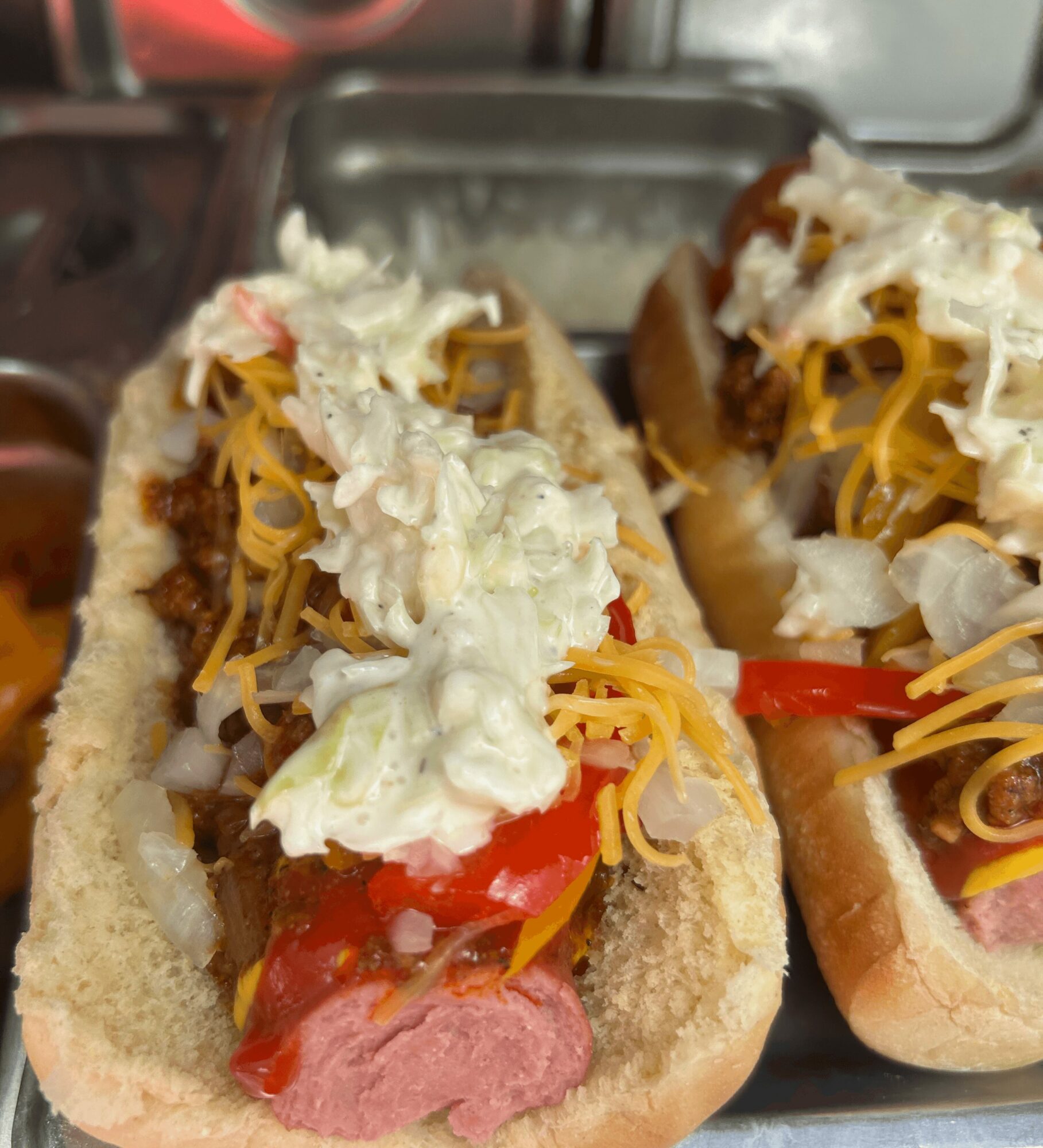 Two hot dogs in buns topped with shredded cheese, chopped lettuce, tomato slices, and mayonnaise, on a tray.