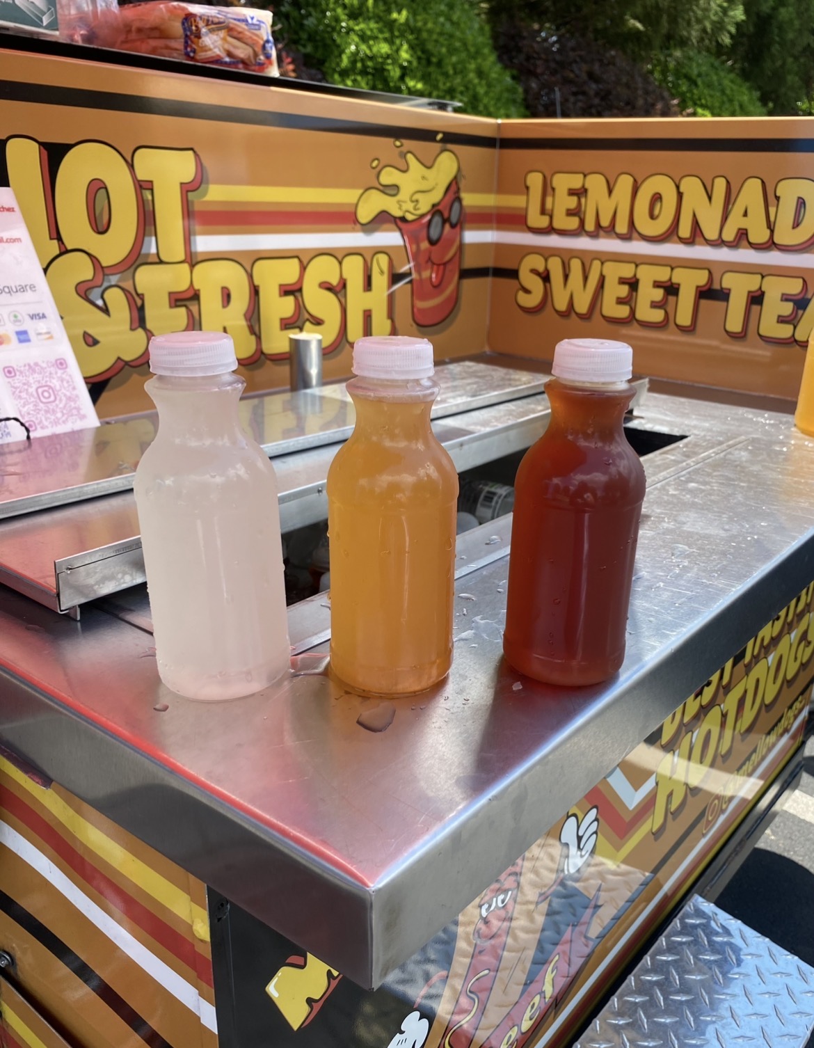 Three bottles of lemonade and sweet tea on a metal counter with a colorful sign in the background.