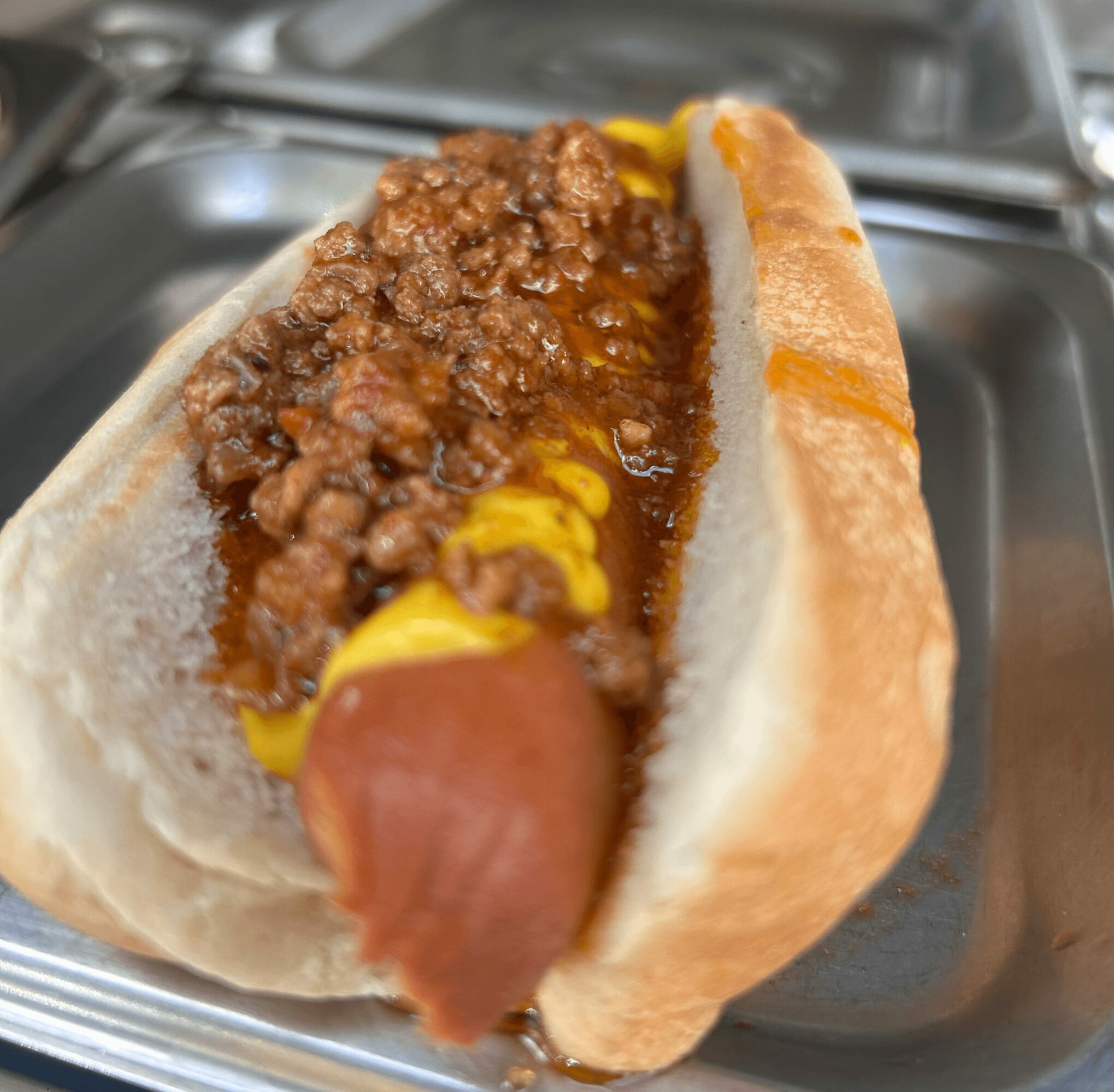 Hot dog with ground meat, mustard, and ketchup in a bun on a metal tray.