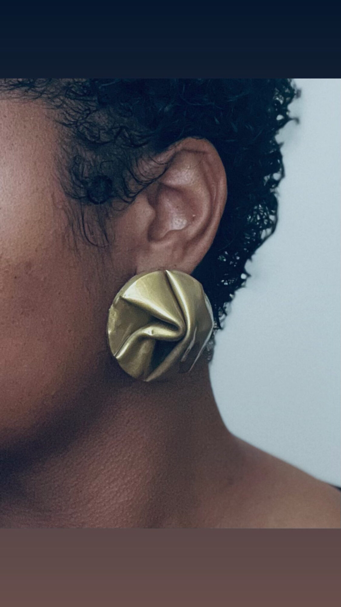 Close-up of a person's ear with a large, gold, twisted earring, and short curly hair, against a light background.