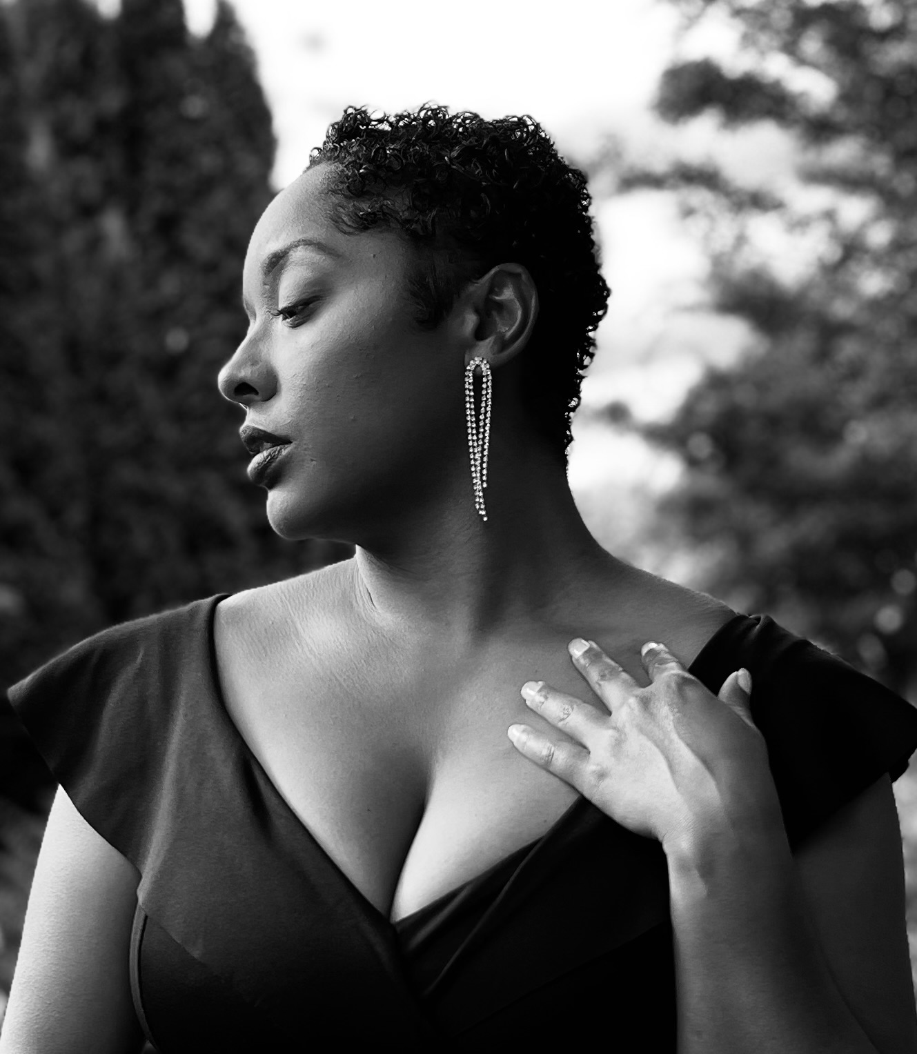Woman with short curly hair wearing a dress and long earring, outdoors, touching her shoulder, in black and white.